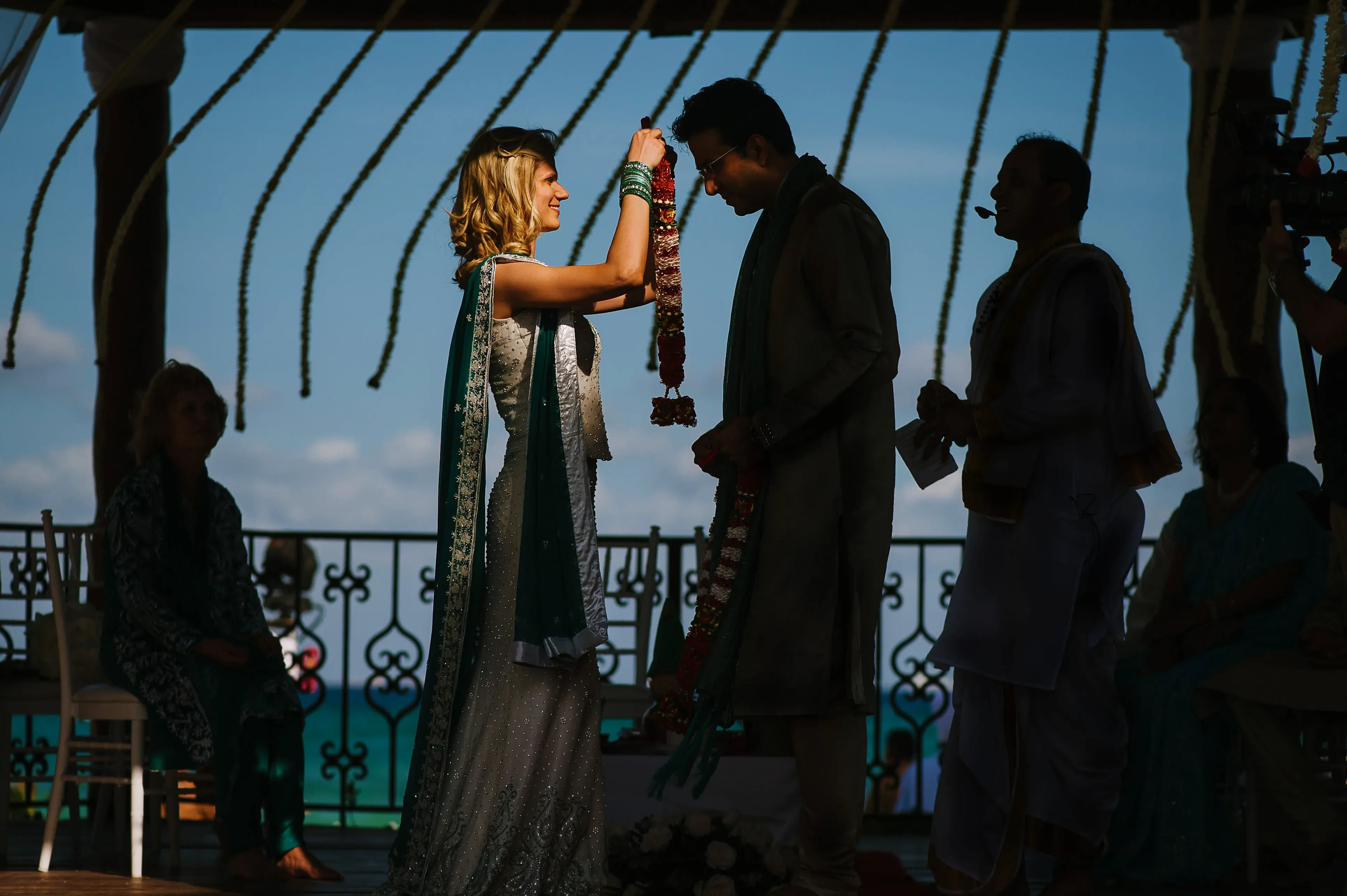 Photo By Citlalli Rico. A bride in a white and green lehenga places a floral garland around the groom's neck during a traditional Hindu wedding ceremony on a balcony overlooking the ocean.