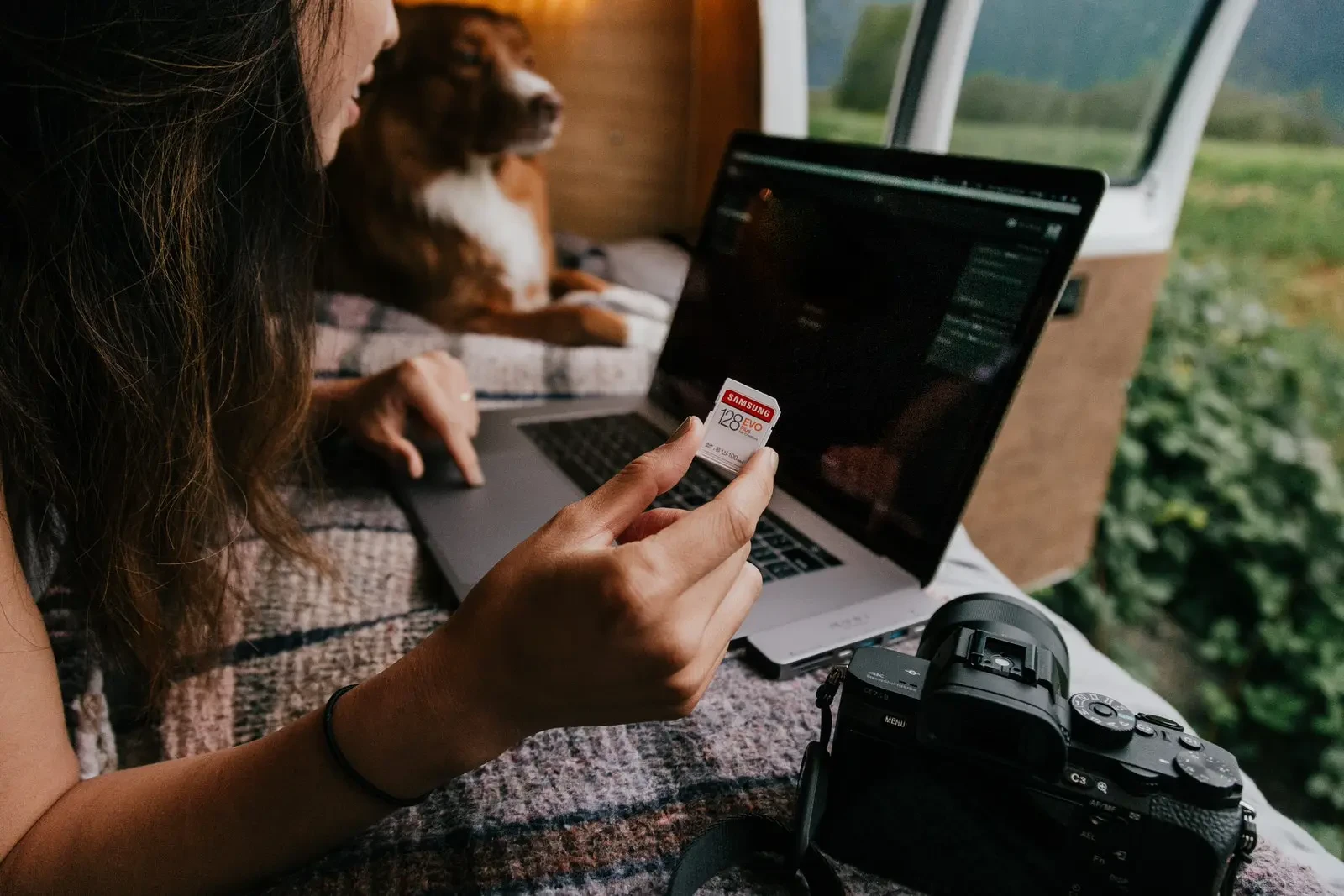 A woman holding a Samsung 128GB EVO Plus SD card while working on a laptop inside a van, with a professional camera and a dog in the background.