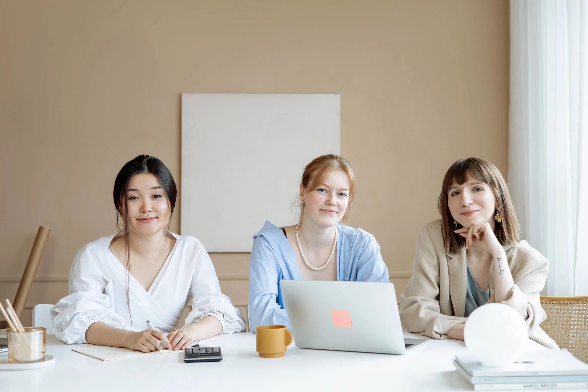 Three businesswomen collaborating at a desk with a laptop in a modern office setting. Photo by Artem Podrez