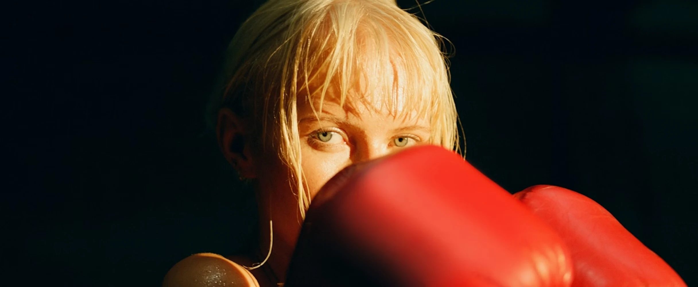 Close-up of a blonde boxer partly hidden behind bright red boxing gloves, staring intensely toward the camera in warm, dramatic light against a dark background.