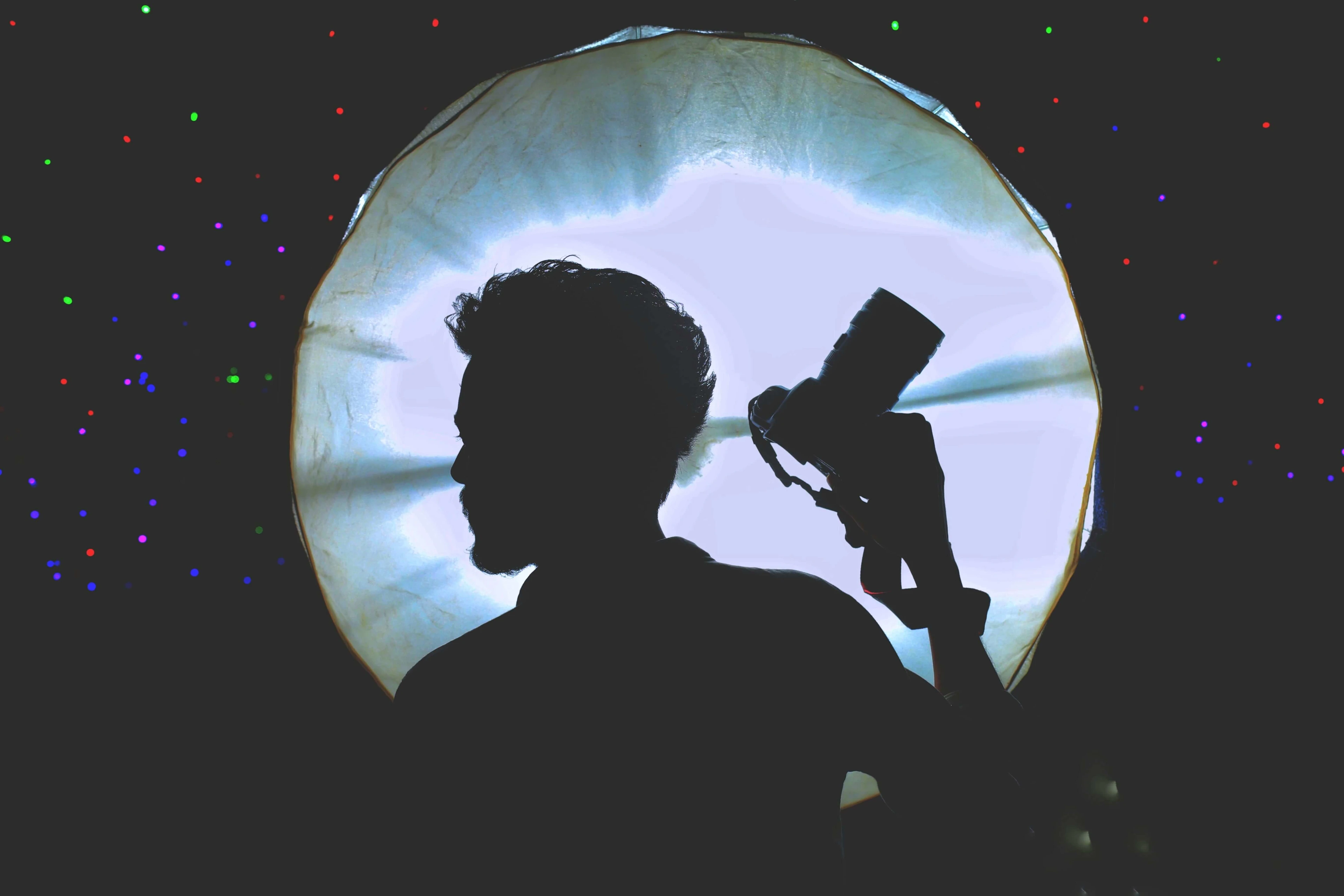 A creative silhouette of a person holding a camera against a bright light source with colorful spots in the background. Photo by SR  Raju: https://www.pexels.com/photo/silhouette-of-person-with-camera-against-light-36893949/