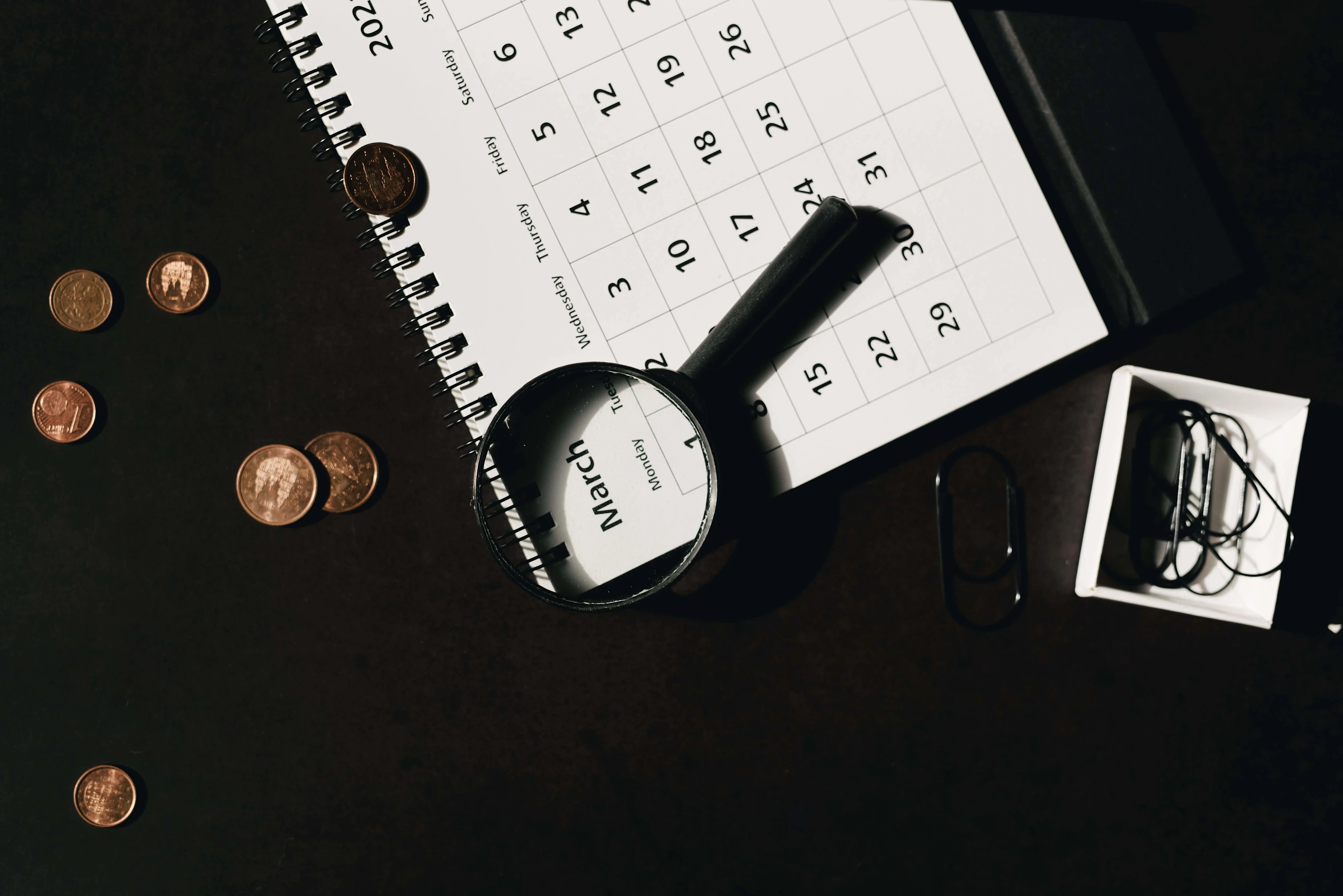 Top view of a calendar with a magnifying glass and coins, ideal for financial themes. Photo by Leeloo The First.