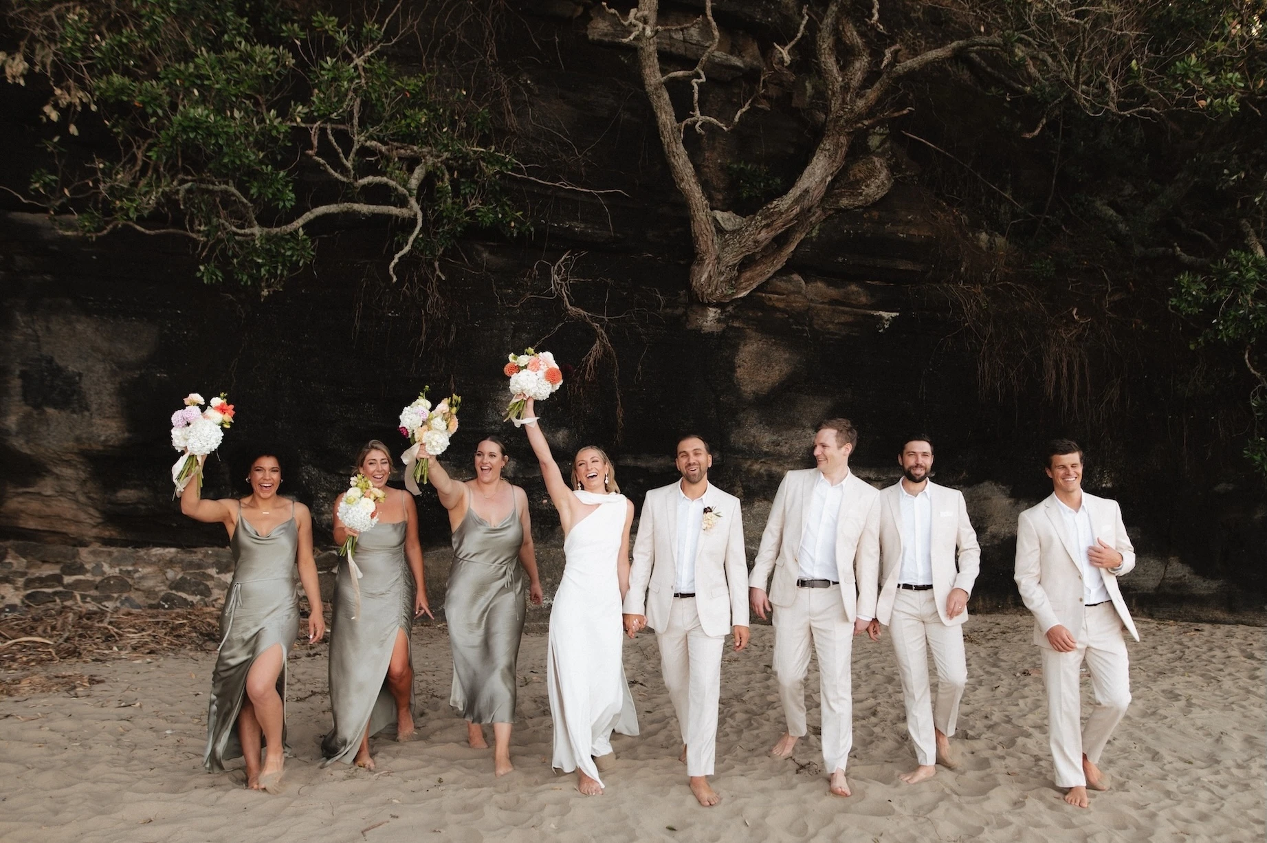 A joyful wedding party walking on a sandy beach, with bridesmaids in olive green dresses and groomsmen in tan suits cheering. Photo By Jonathan Suckling