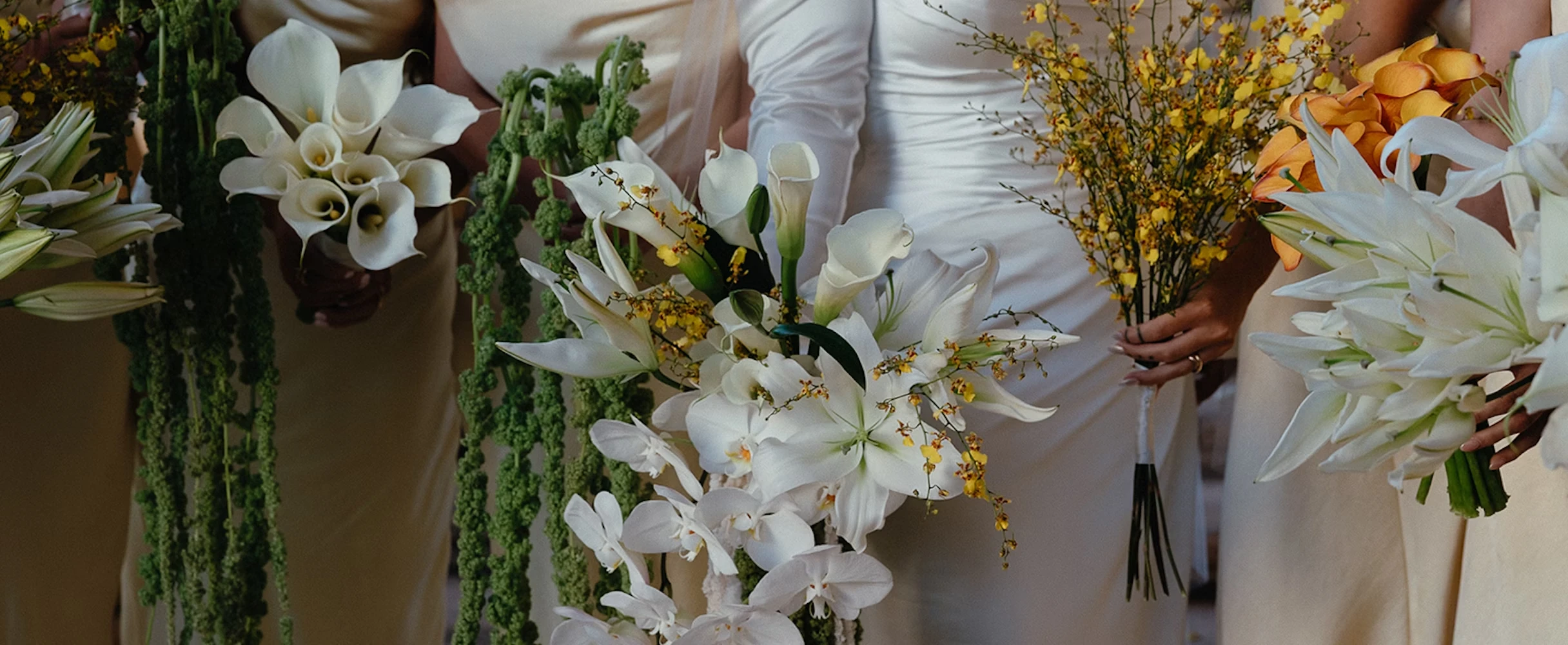 Bridal party in white dresses holding elegant bouquets of white calla lilies, orchids, and yellow accent flowers at a wedding ceremony