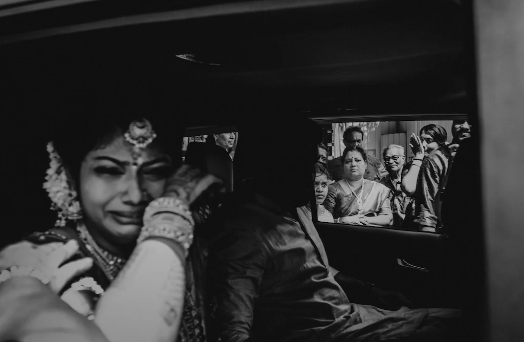 Photo By Joyful Project. A bride in her wedding dress sits in the back seat of a car, in tears.