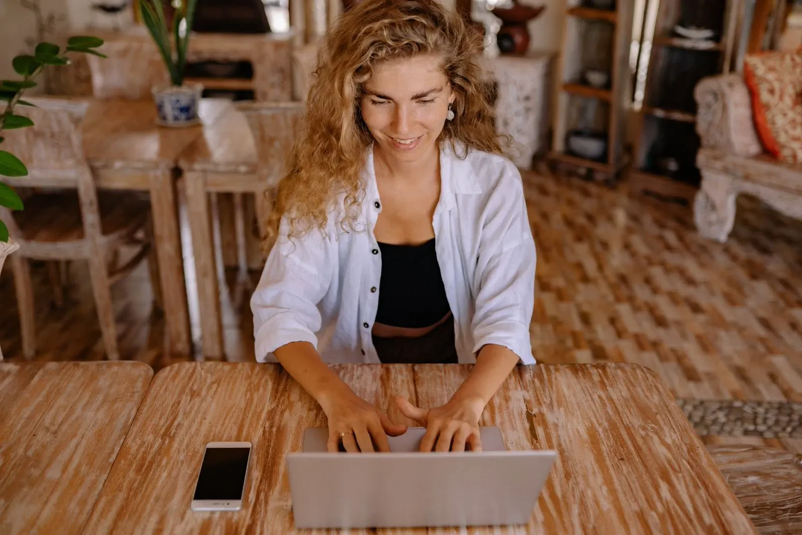 A woman with curly hair smiling while typing on a laptop at a rustic wooden table in a brightly lit cafe.