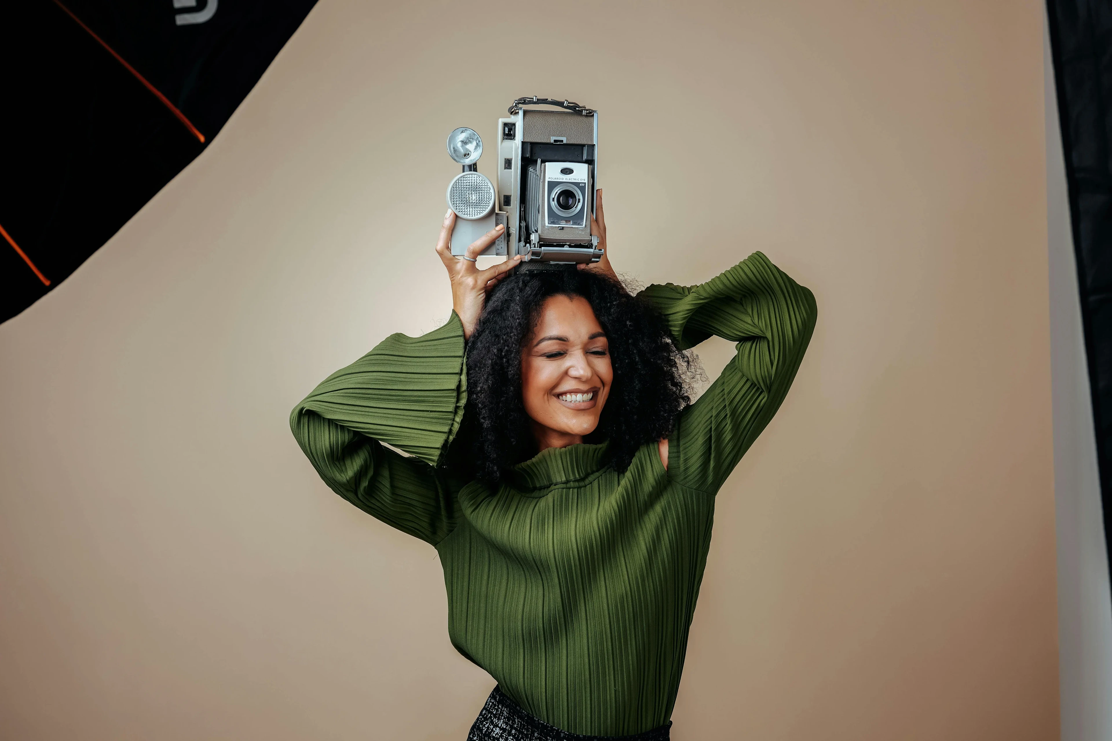 An image of a smiling woman with dark curly hair wearing a green ribbed sweater, playfully balancing a vintage Polaroid Land Camera on her head against a beige studio backdrop. Photo by Pexels LATAM: https://www.pexels.com/photo/photo-of-a-brunette-woman-holding-a-vintage-camera-above-her-head-16135549/