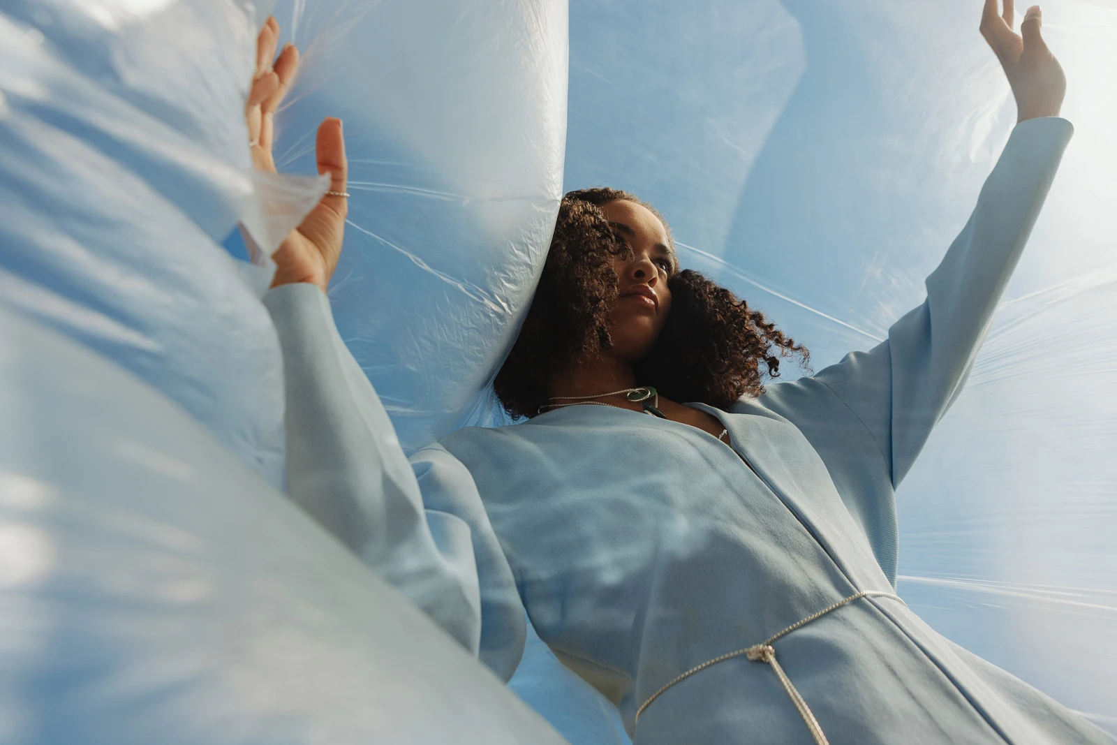 Photo by Camila Berrio. Low-angle fashion portrait of a woman with curly hair wearing a light blue dress, posing behind layers of translucent plastic sheeting against a clear blue sky.