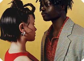 A portrait of a man and a woman looking at each other against a bright yellow background. The woman has dark braided hair, a red top, and large silver hoop earrings; the man has dreadlocks and wears a patterned red shirt under a light blazer.
