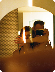 A couple taking a mirror selfie in a bathroom. A woman with reddish-brown hair partially covers her face with her hand while a man behind her holds a vintage camera.