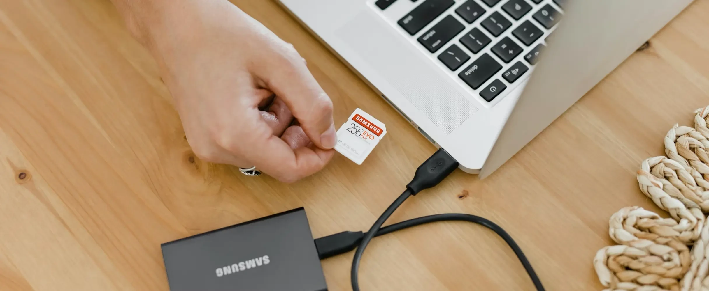 A person's hand holding a Samsung 256GB EVO Plus microSD card next to a silver laptop and a Samsung external portable SSD on a wooden desk.