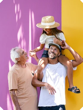 A multi-generational family portrait: a younger man smiling with a young girl on his shoulders, and an older man beside them, against a two-tone purple and yellow background with sunlight and soft shadows.