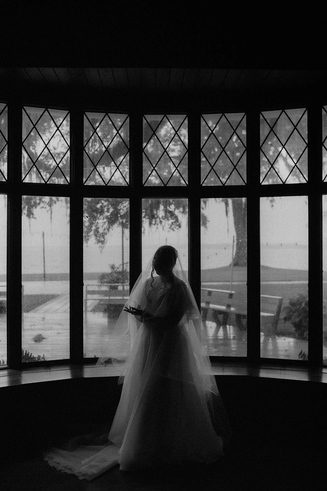 A black and white photo of a bride capturing the light outside the window and silhouetting the bride with only light through her veil. Photo by Milly Baine of the Rose Reflective.