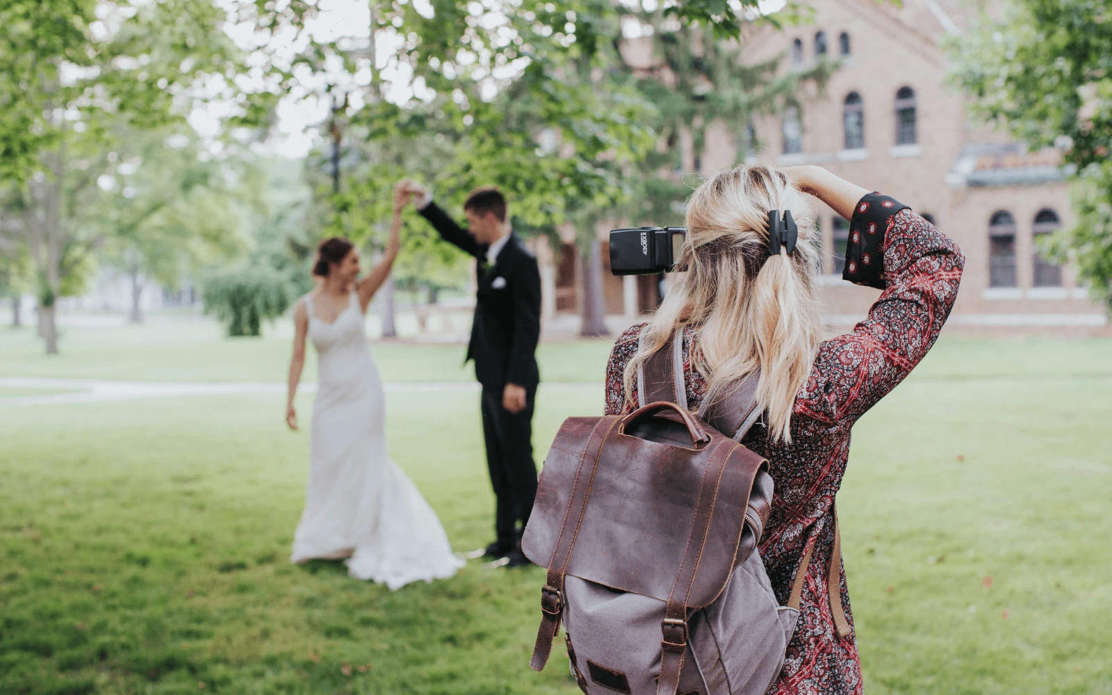 A female photographer wearing a backpack photographs a bride and groom dancing on a green lawn outdoors. Image provided by Peakto.