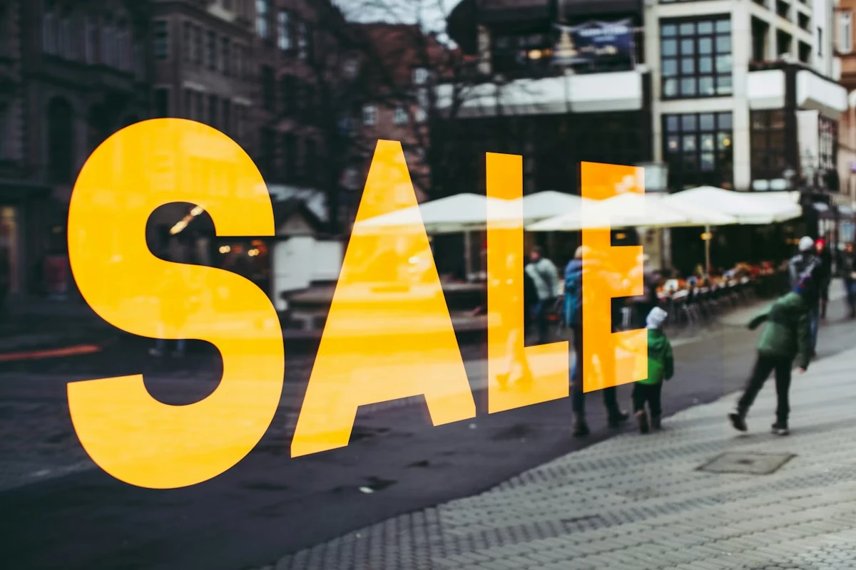 Street scene reflected in a shop window with the word “SALE” in large yellow letters over a city sidewalk with people walking by.