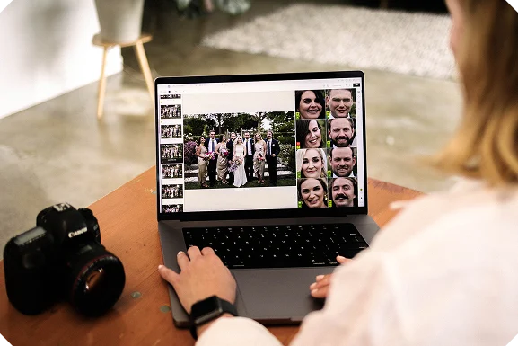 A person seen from behind at a wooden table with a silver laptop and a black DSLR beside it. The screen shows a photo editing interface with a grid of six faces on the right and a larger group photo, possibly a wedding, on the left.