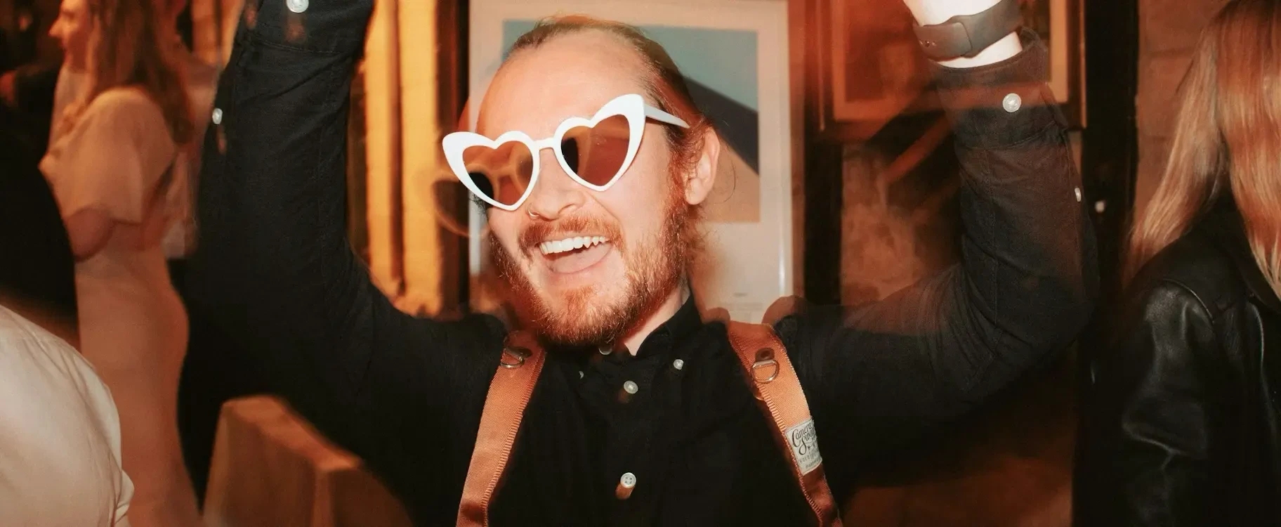 Dancing and Dive-Rolling with Wedding Photographer Jonathan Suckling. A cheerful man with a beard and white heart-shaped sunglasses laughing with his arms raised at a dimly lit event.