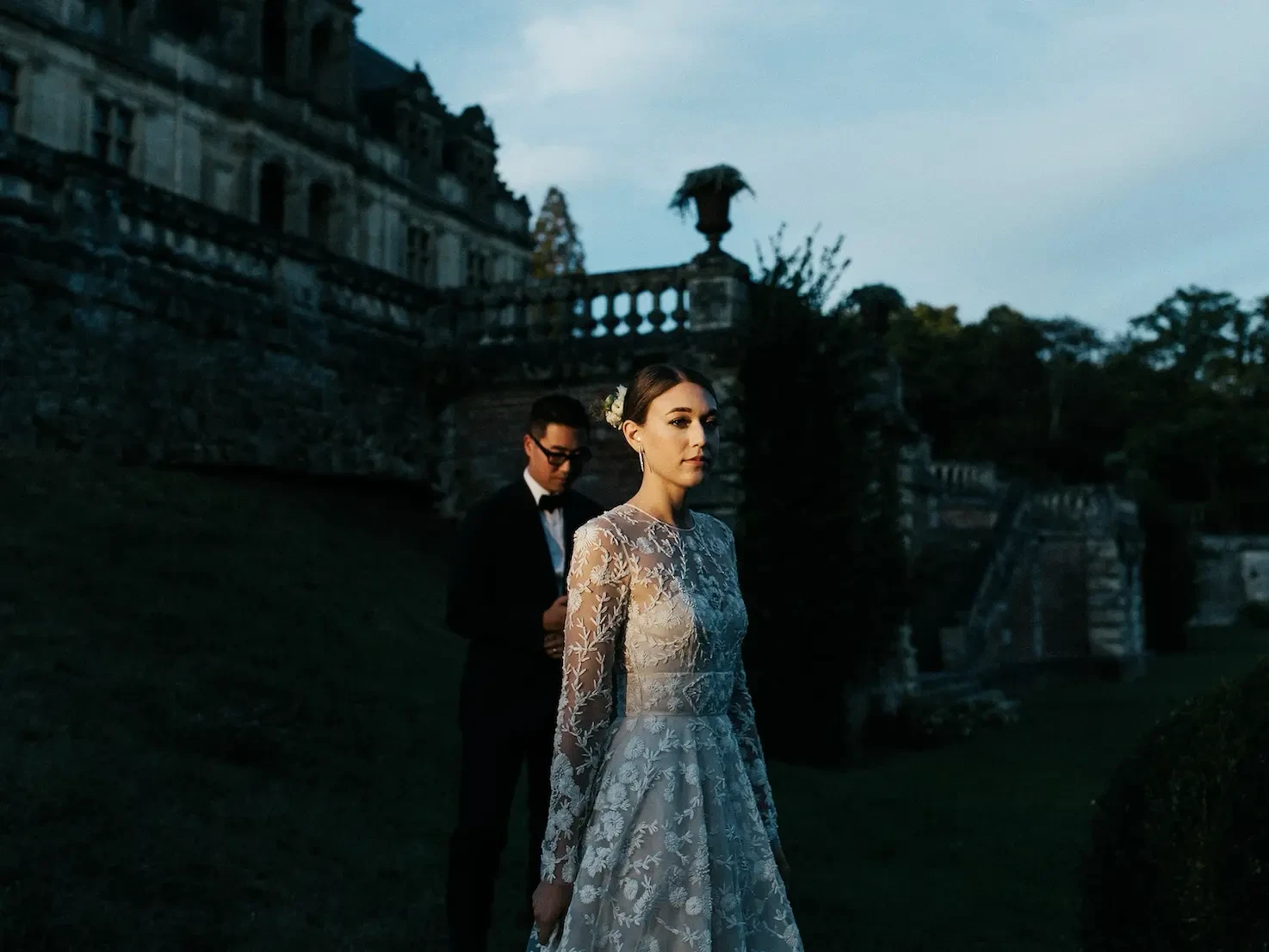 Photo By Ryan Browne. A bride in an intricate lace long-sleeve gown stands in soft sunlight in front of a grand, dark stone European-style estate at dusk.