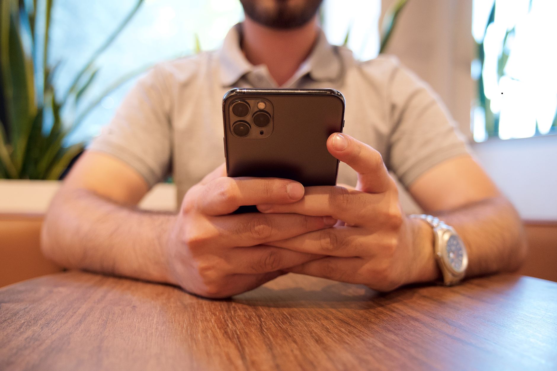 Ein Mann hält ein Smartphone an einem Cafétisch; Pflanzen sind im Hintergrund zu sehen · Foto: iam hogir (Pexels)