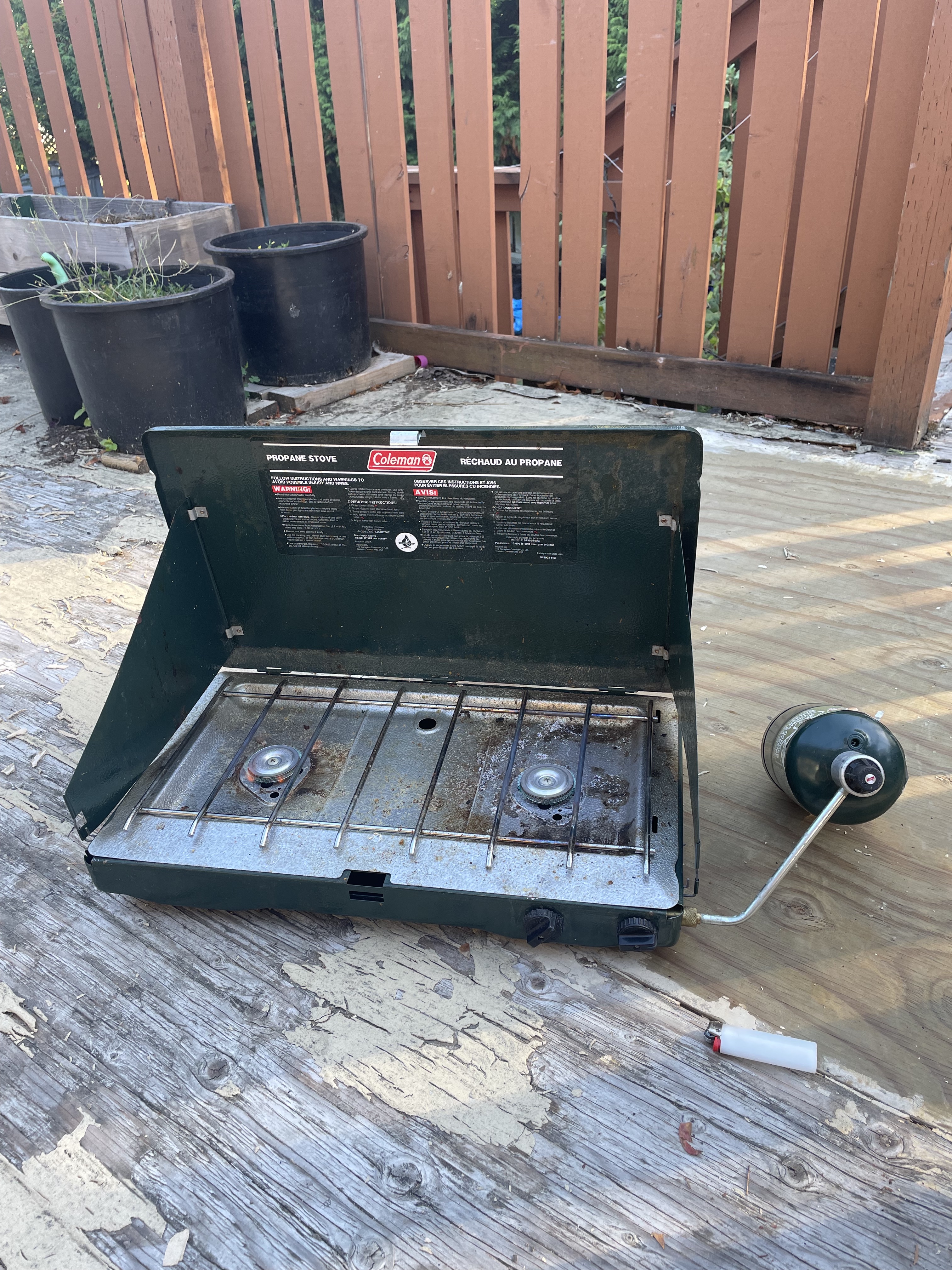 A picture of a green open camping stove, with two burners covered by a metal grill with wide gaps between the bars, and a propane canister attached.