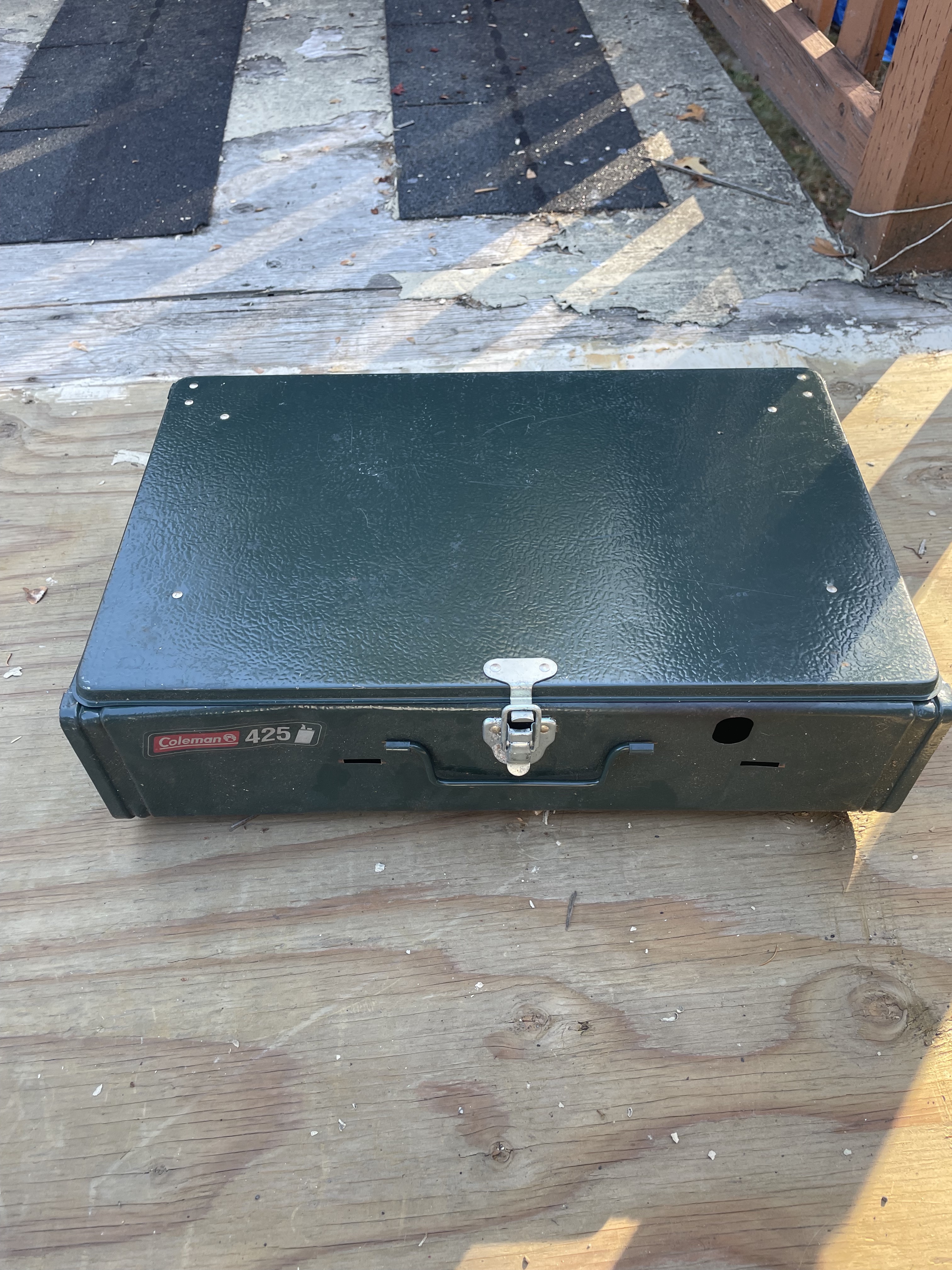 A rectangular and heavy looking metal double burner liquid fuel stove closed and ready for transport. It has a locking buckle and a thin handle for carrying.