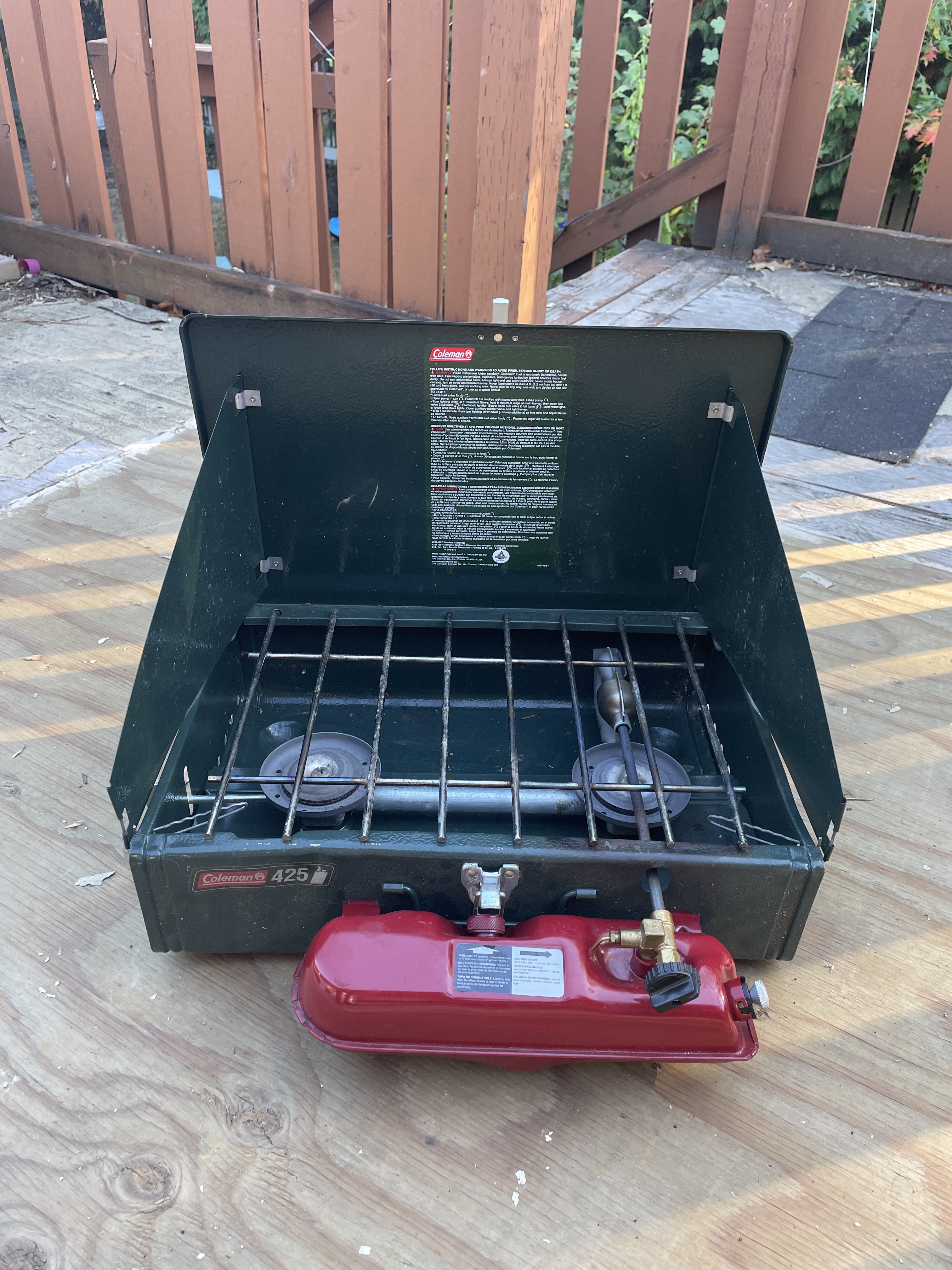 A picture of a Coleman double burner stove with metal grates over the two burners. A refillable fuel container is attached to the front.