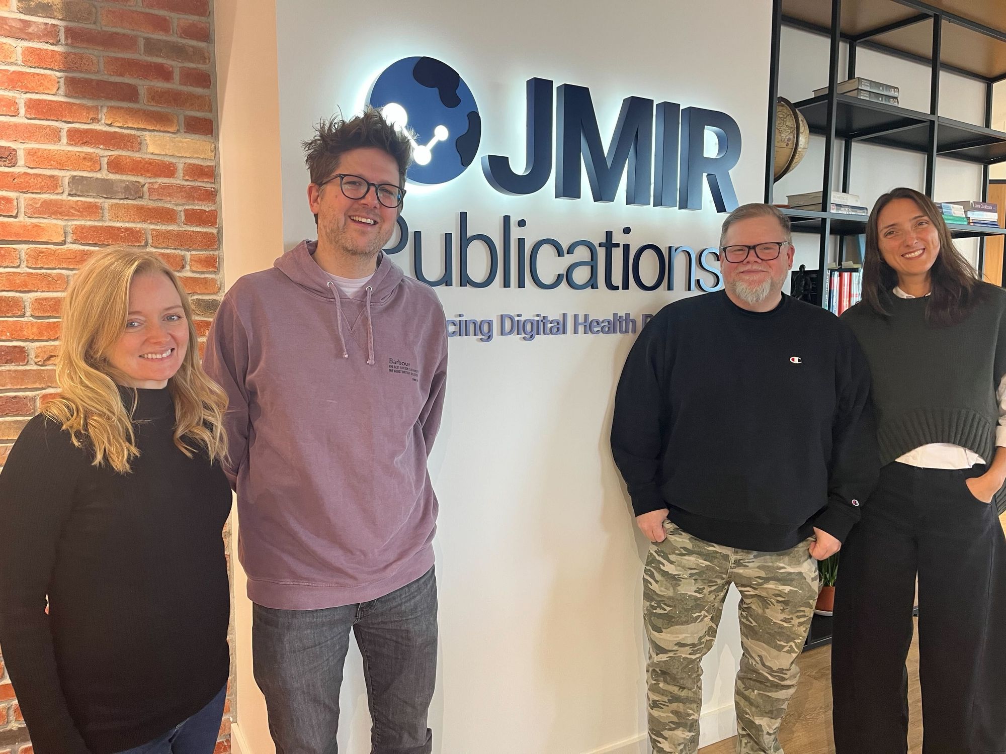 A photo of some members of the PREreview team in front of the JMIR Publications Logo. From left to right, Vanessa Fairhurst, Chris Wilkinson, Chad Sansing, and Daniela Saderi.