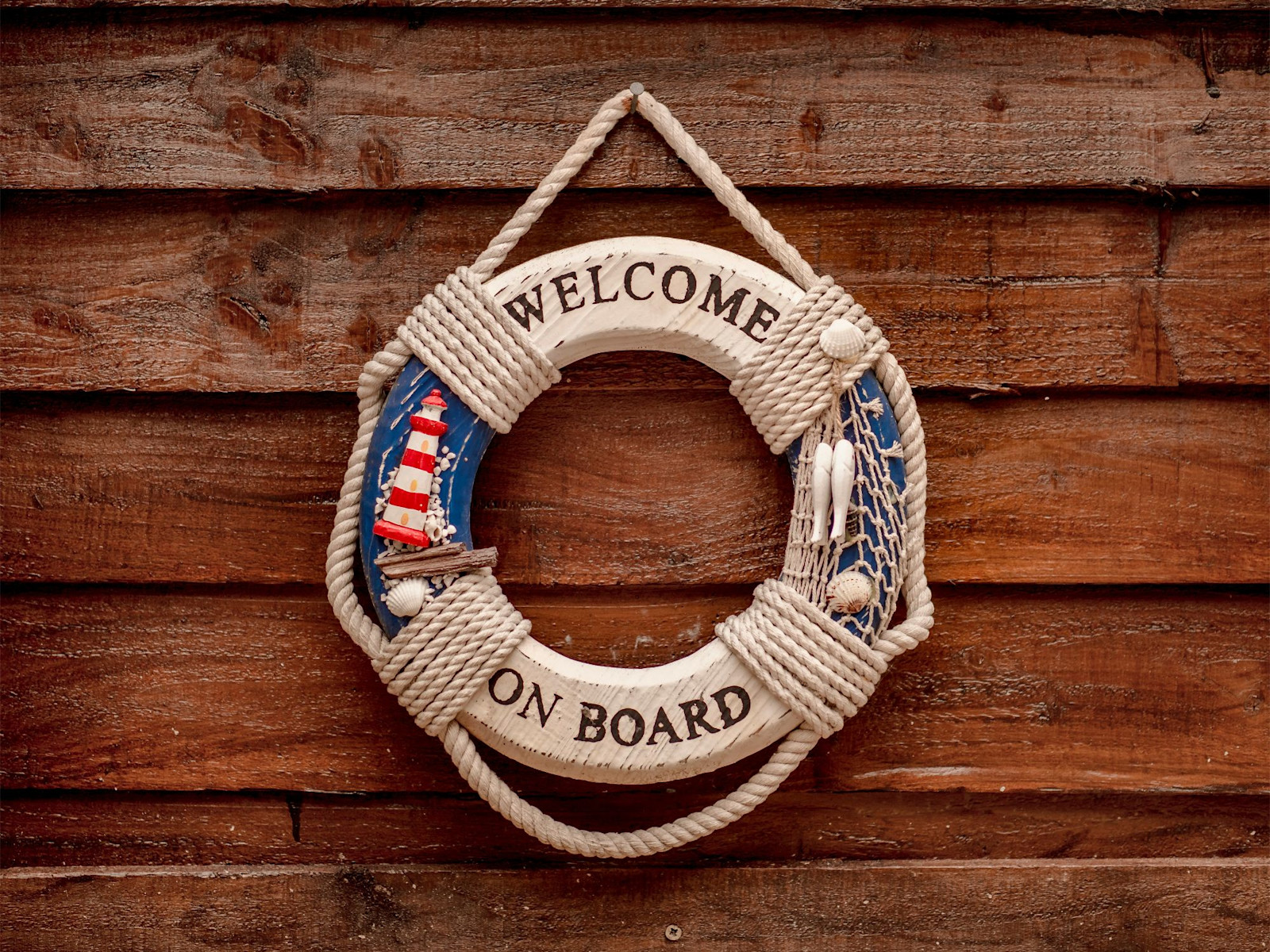 Nautical-themed lifebuoy with "Welcome On Board" message hangs on rustic wooden wall.