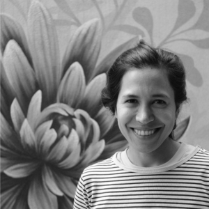 Black and white headshot of María Sol Ruiz (she/her), smiling and wearing a striped shirt and tied up long hair.
