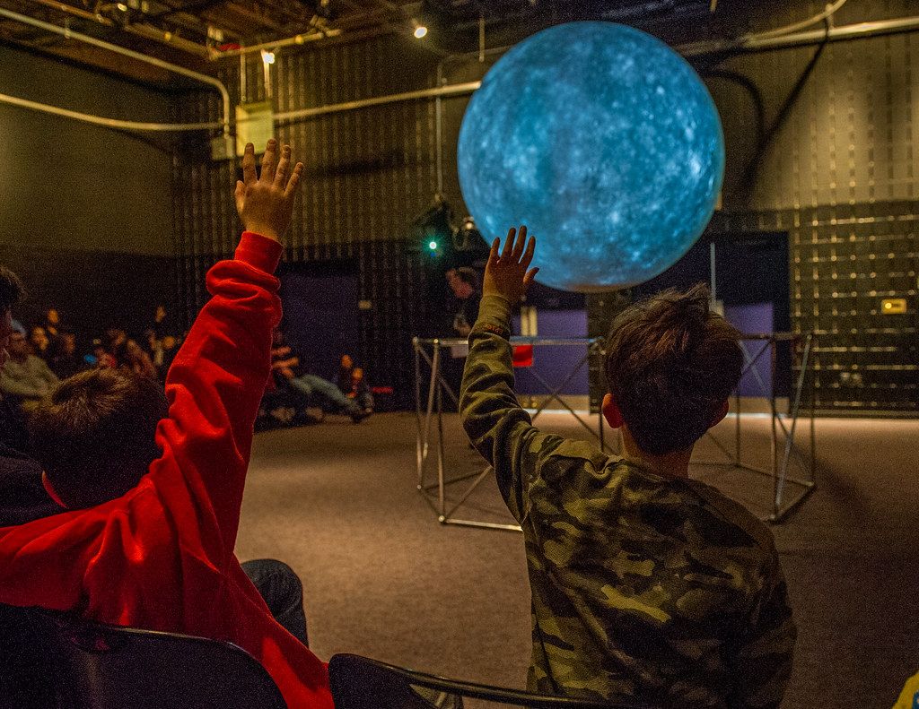 Color photograph of two children seen from the back who are raising their hand toward a glowing globe at what is presumably a NASA-organized event. More people in the background.