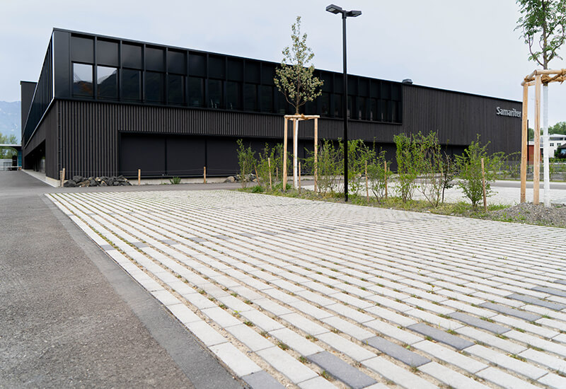 Ansicht eines Parkplatzes vor einem modernen, dunklen Gebäude mit hellen Ökosteinen, die durch breite Fugen eine Versickerung von Regenwasser ermöglichen.