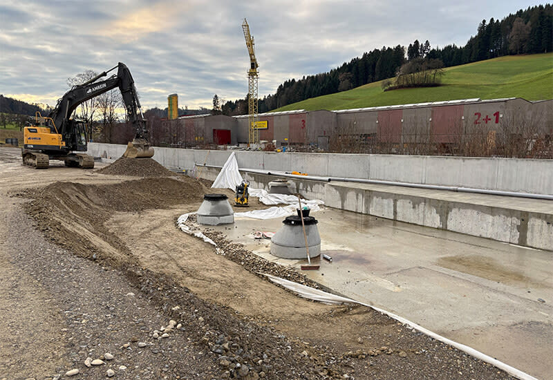Blick auf Bauarbeiten für das neue Kieswerk Sandacher auf einem Werksgelände mit Bagger, Betonschächten und einer Industriehalle vor einem Waldhang.