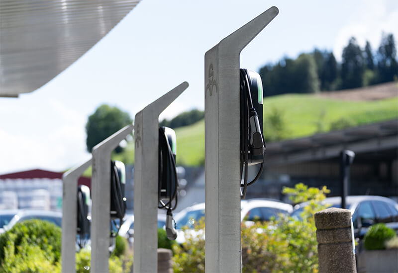 Reihe von Elektroladesäulen aus grauem Beton mit montierten Wallboxen auf einem Parkplatz vor grüner Hügelkulisse.