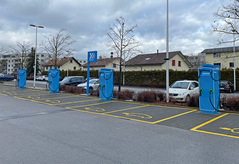 Eine Reihe von vier blauen GOFAST-Schnellladestationen auf einem gepflasterten Parkplatz unter bewölktem Himmel.
