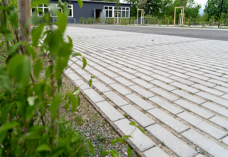 Vue en perspective d'un parking recouvert de pavés écologiques perméables, avec au premier plan les feuilles vertes d'une haie et, à l'arrière-plan, un bâtiment d'entreprise moderne.