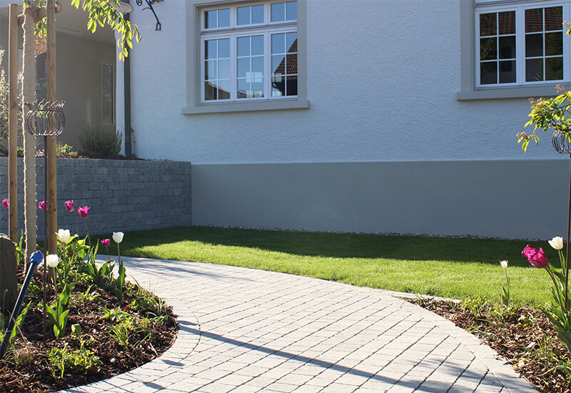 Gartenplatz mit einem Weg aus Betonpflastersteinen, einer Hausfront und einer Mauer aus Betonsteinen. Eingerahmt durch eine grüne Wiese und farbige Tulpen. 