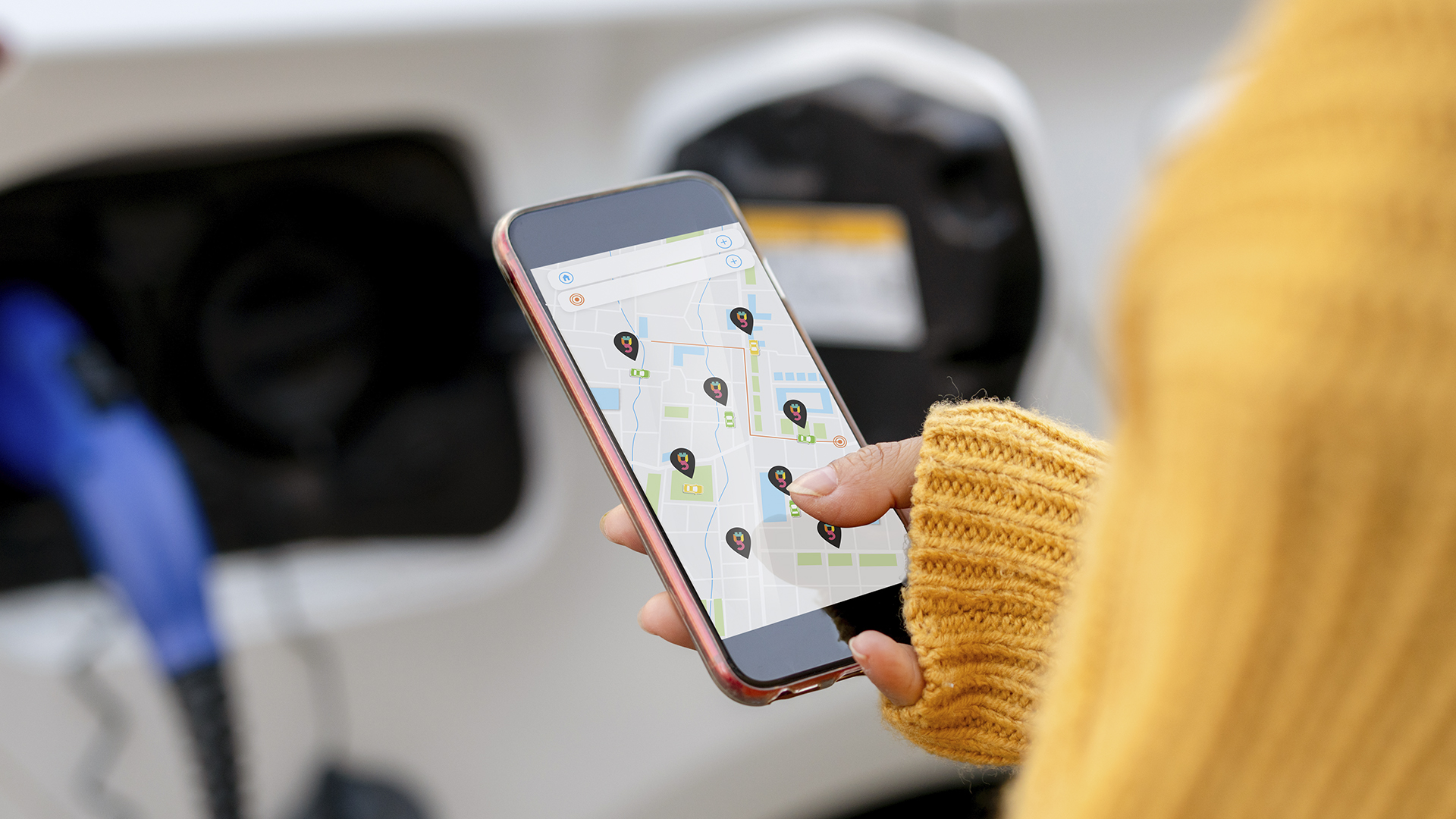 In the background, a car charging port is being used. In the focus of the image, a mobile phone shows a map with several pins on it in various locations, indicating several charging points.
