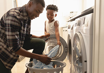 Smart Hints and Tips: Laundry IE HE A man and a child adding freshly washed clothes from a washing machine into a laundry basket