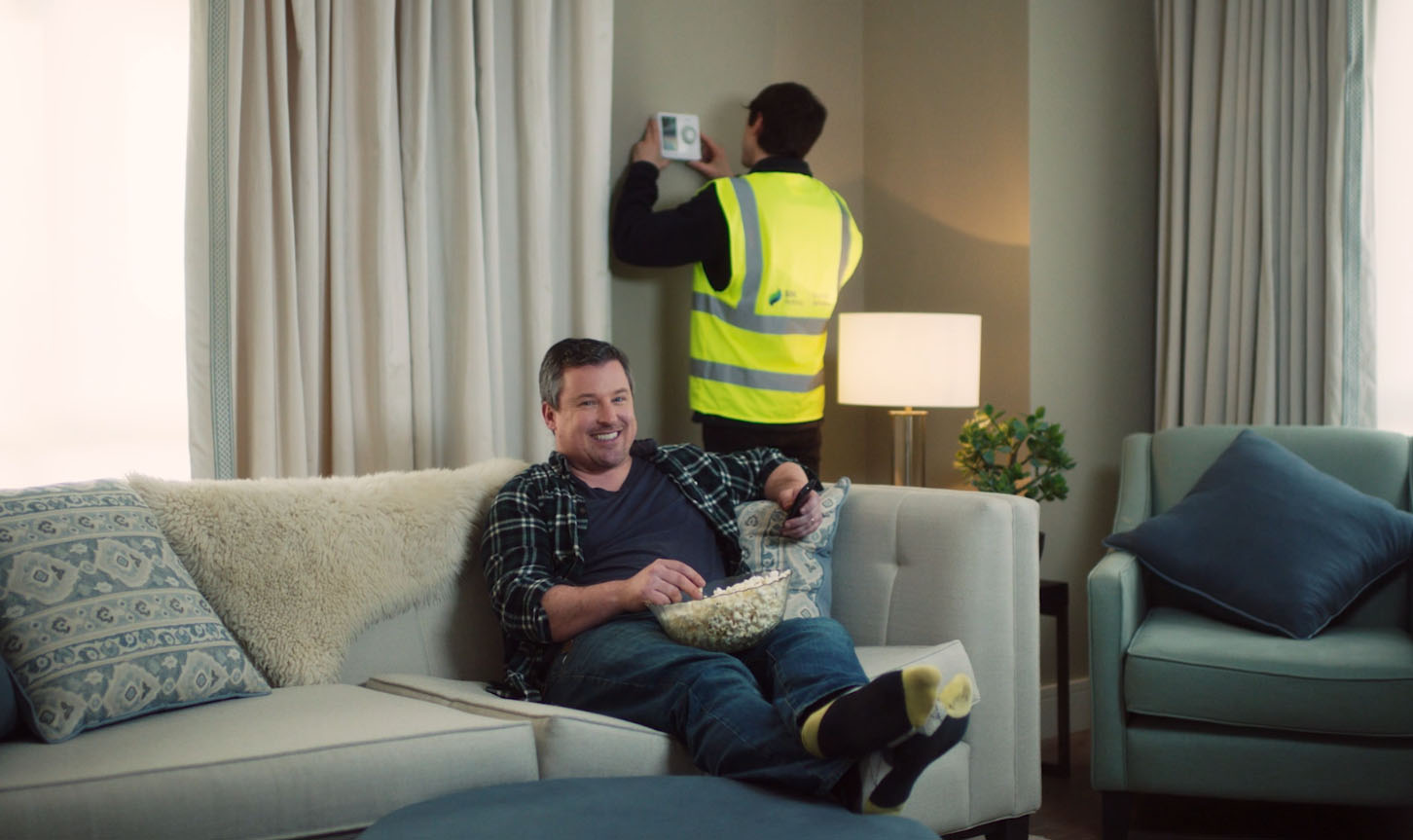 A man sitting on a sofa holding a tv remote and holding a bowl of popcorn while someone from SSE work on the themostat