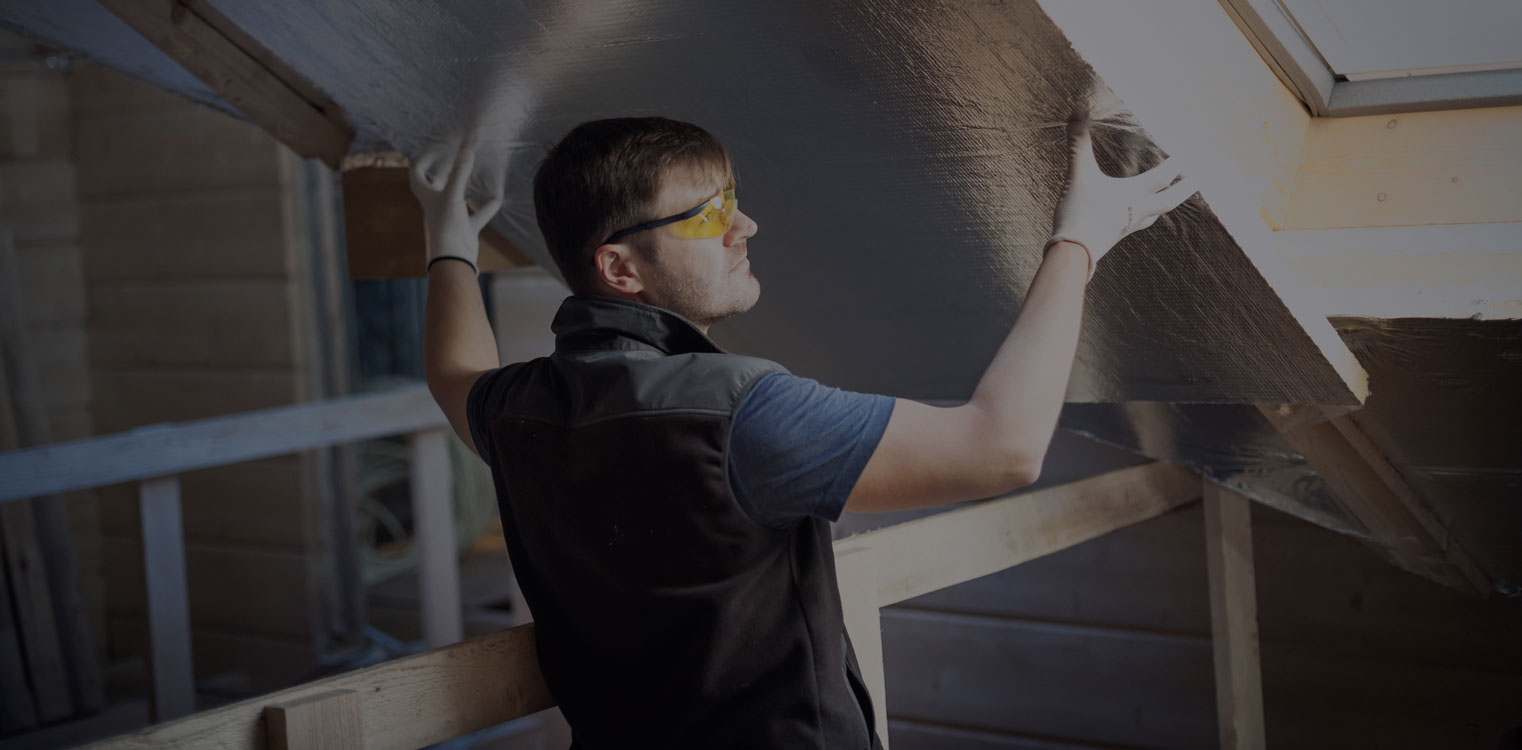 Insulation-hero (overlay) A man wearing a dark vest, light-colored shirt, safety glasses, and gloves working in an attic