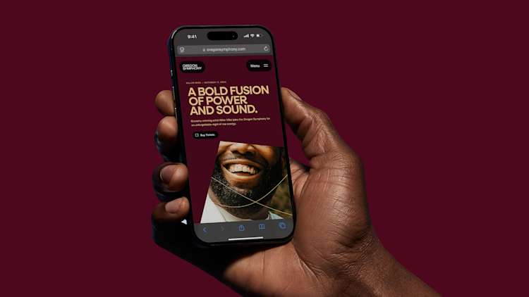 A hand holds an iPhone displaying the Oregon Symphony website against a burgundy background. The screen shows a promotional design with golden text reading 'A BOLD FUSION OF POWER AND SOUND' above a close-up photograph of a bright smile. A 'Buy Tickets' button is visible below the text.