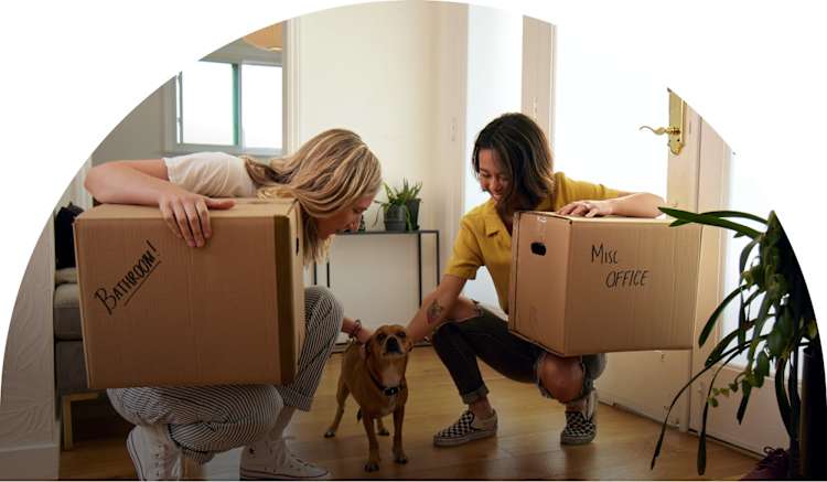 Two women carrying moving boxes and petting a small dog.