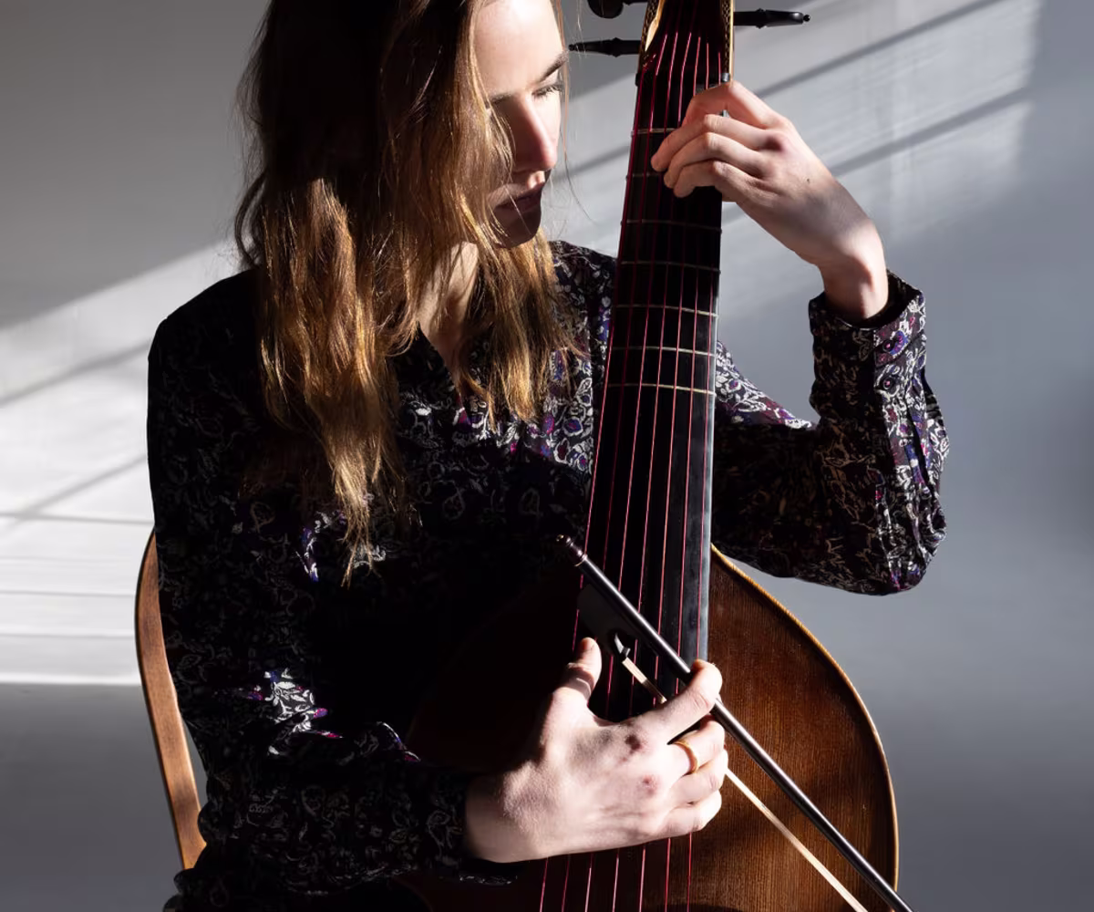 Good Again? A woman, sarah small, playing the viola da gamba pizzicato with rays of light illuminating her face and hands from an adjacent open window