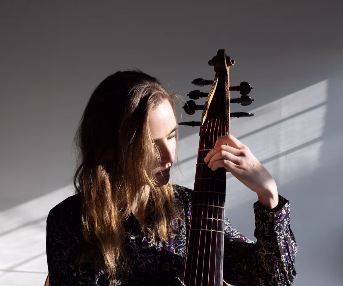 Good Again? A woman, sarah small, playing the viola da gamba pizzicato with rays of light illuminating her face and hands from an adjacent open window