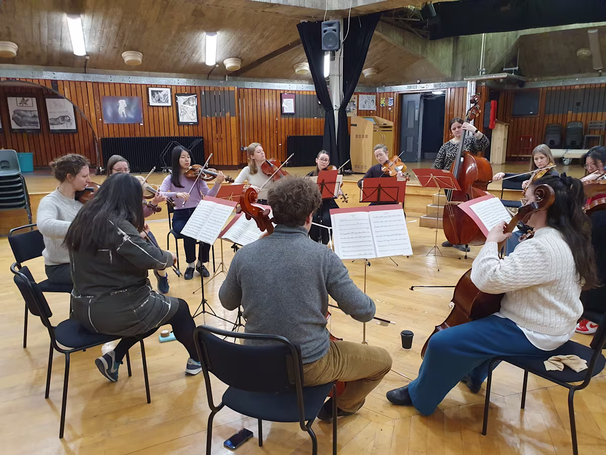 Applications open for prestigious orchestral experience scheme classical musicians sitting in a circle in a large rehearsal space with wooden floors and walls