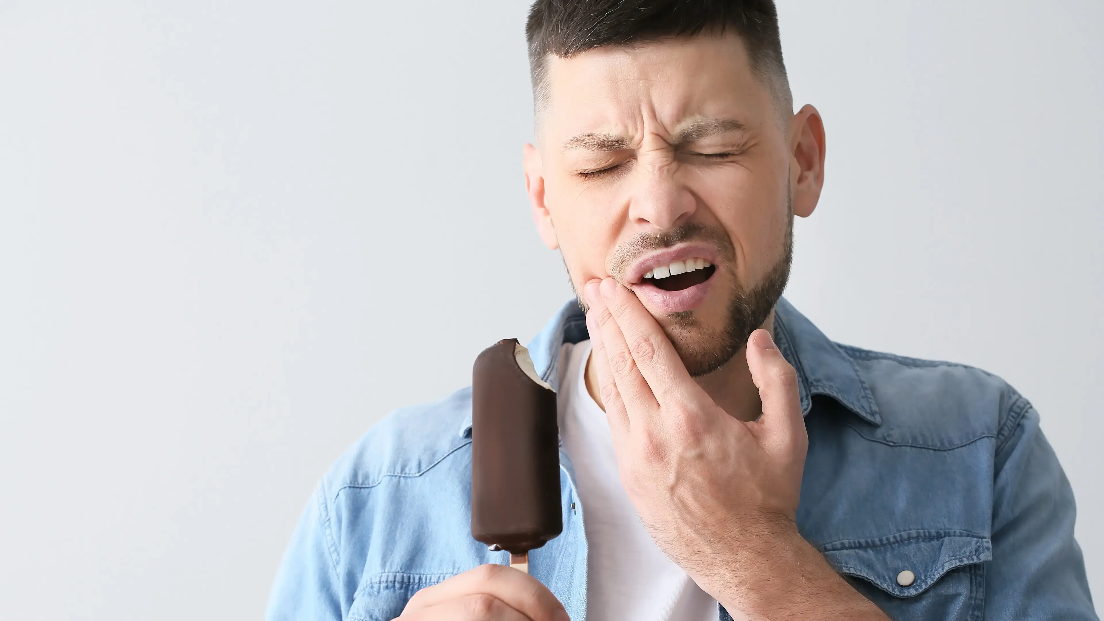 Ein Mann, der ein Eis in den Händen hält und dabei schmerzvoll schaut und seine Hände an der Wange hat.