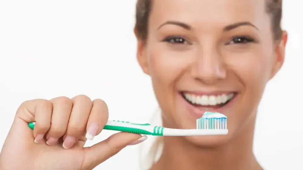 Eine lächelnde Frau mit offenem Mund und einer Zahnbürste mit Zahnpasta in der Hand.​