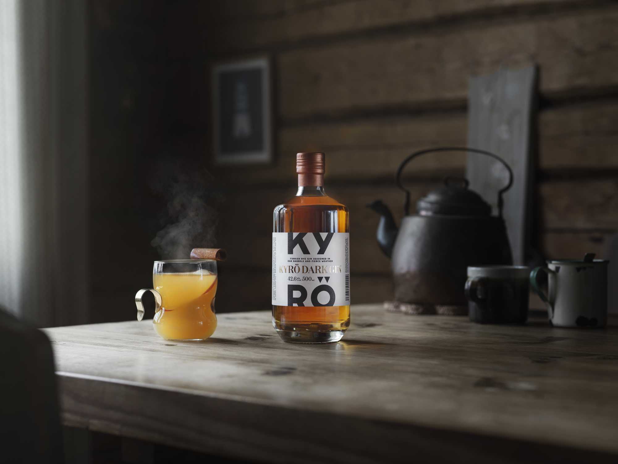A bottle of Kyrö Dark Gin standing on a table in a wooden house, next to it a steaming glass mug filled with hot Dark Gin, apple juice, and a slice of apple. Horizontal. 