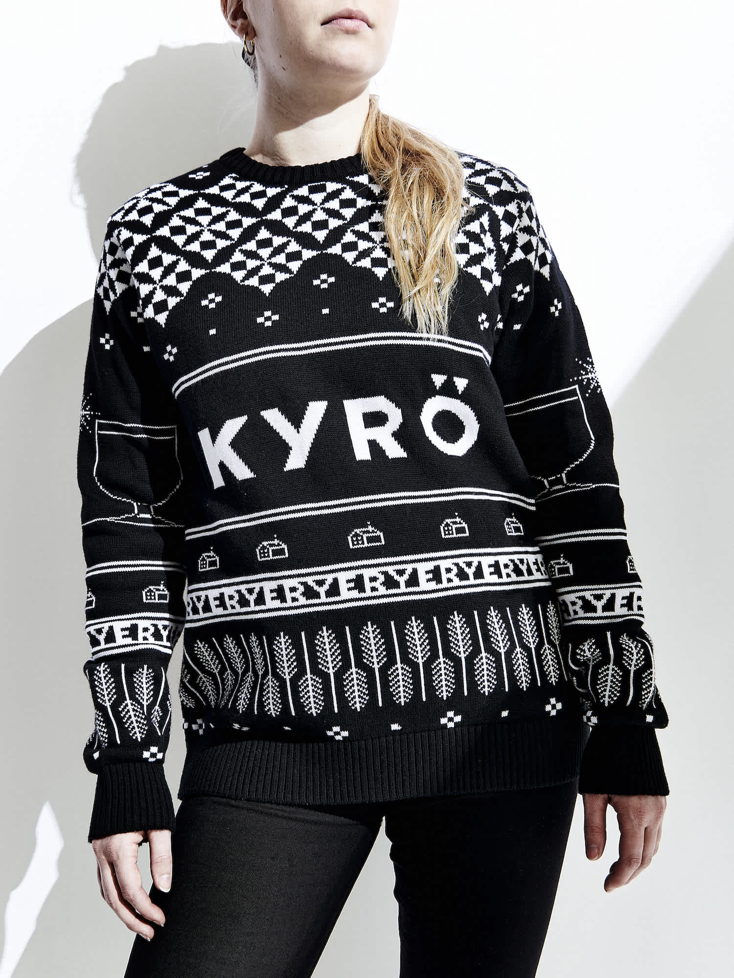 White, female model wearing a black sweater decorated in white, graphic, Christmas illustrations and a big, large Kyrö logo across the chest. 