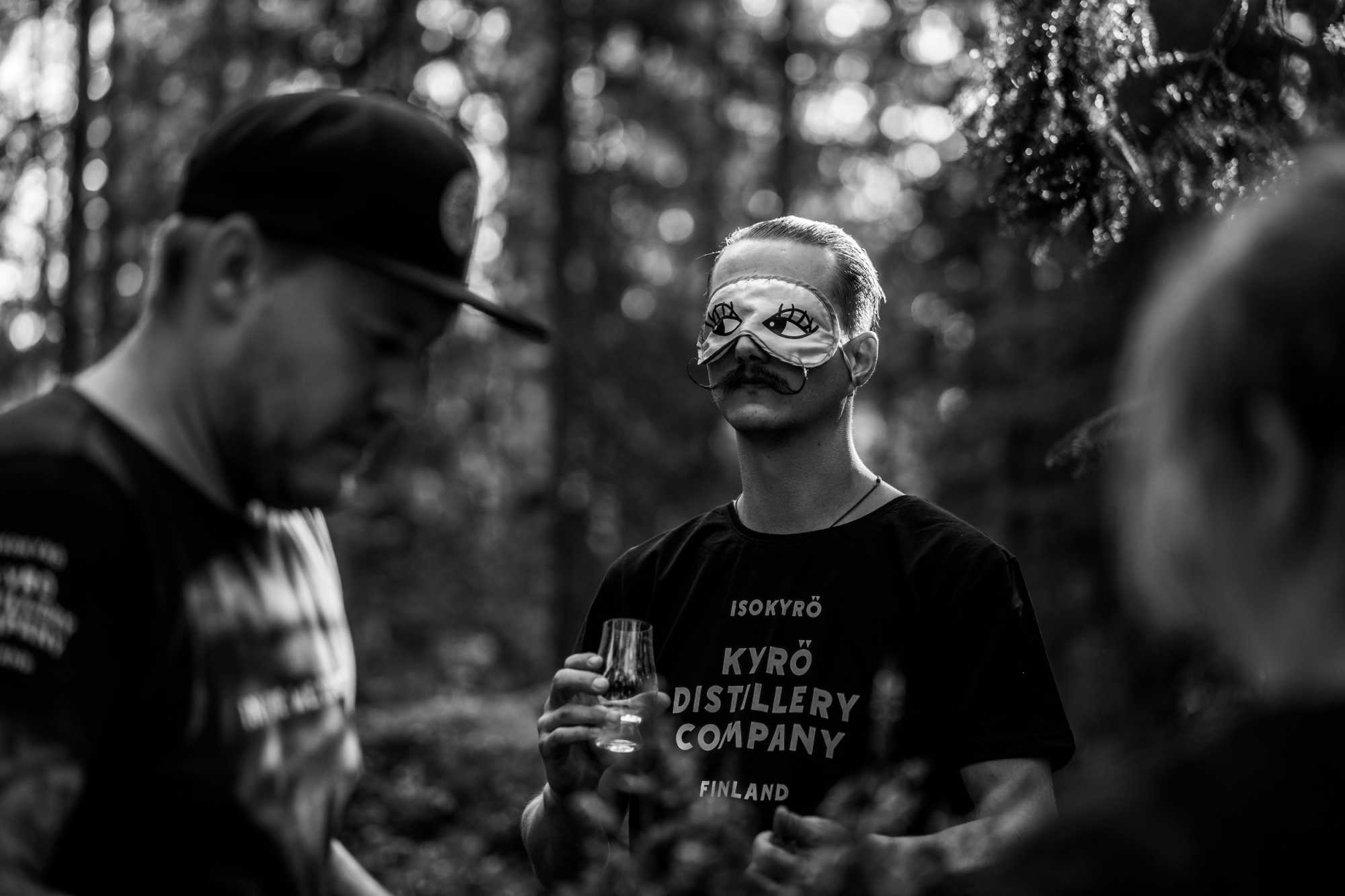 Person with an eye mask holding a whisky tasting glass in a middle of a forest. 